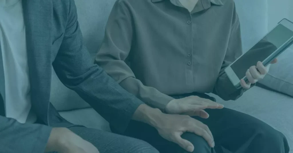 A man’s hand is holding a woman’s leg, as she reaches out her own mentioning him to stop. She is holding a tablet with evidence of sexual harassment in Oregon.