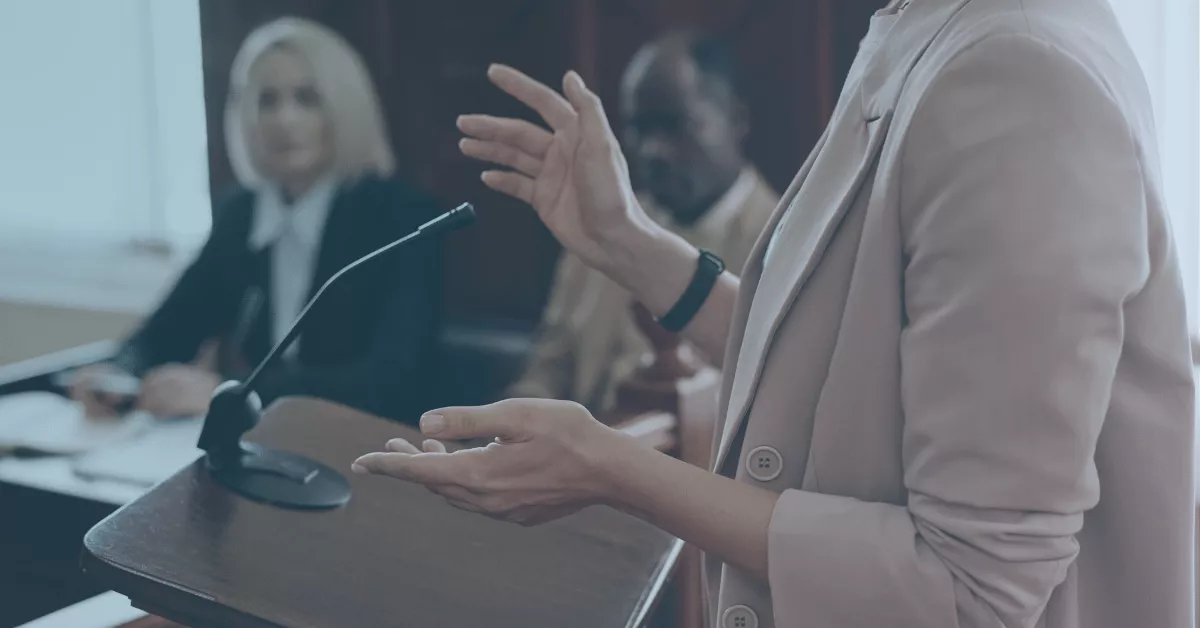 Person speaking at a podium in a courtroom setting, with people seated in the background.