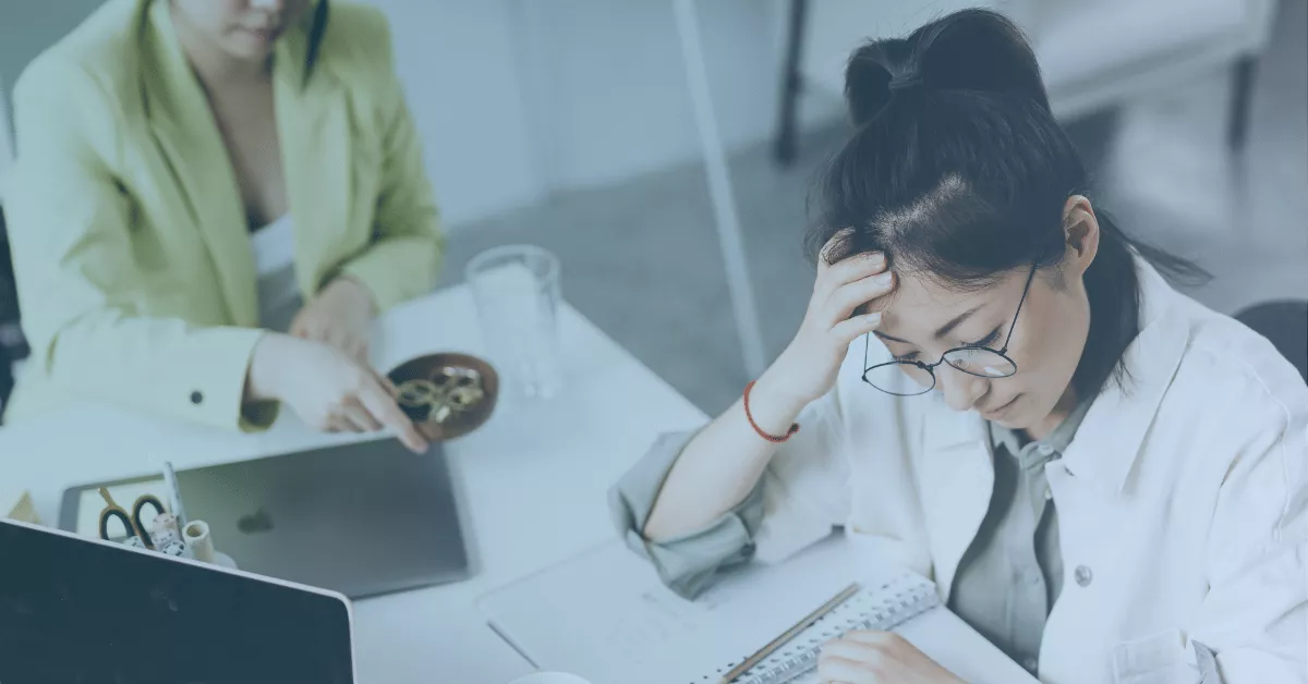 Stressed employee holding their head while looking at notes, with another person across the desk gesturing near a laptop.